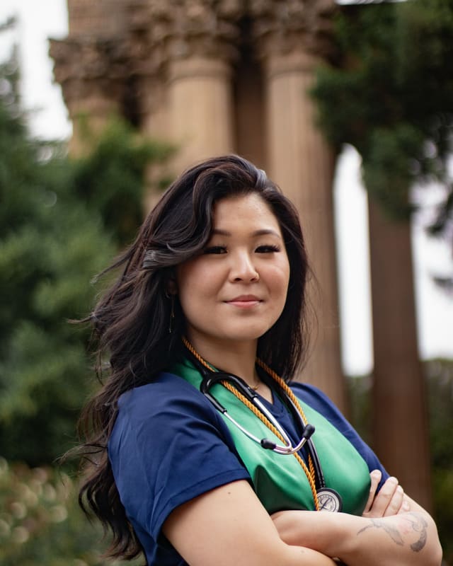 A nursing graduate in scrubs with a stethoscope and graduation cords